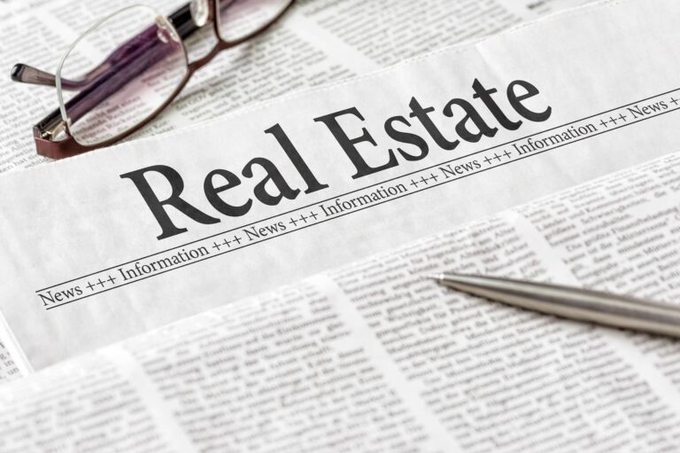 A pair of glasses and a pen resting on a newspaper section titled "real estate".